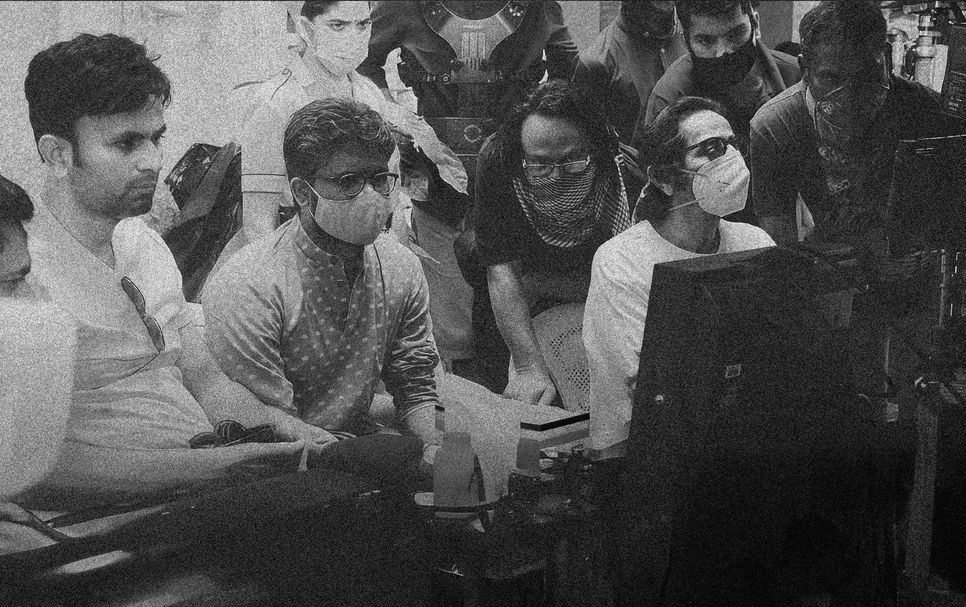 Black-and-white behind-the-scenes photo: crew members, most wearing face masks, watch a monitor on set.