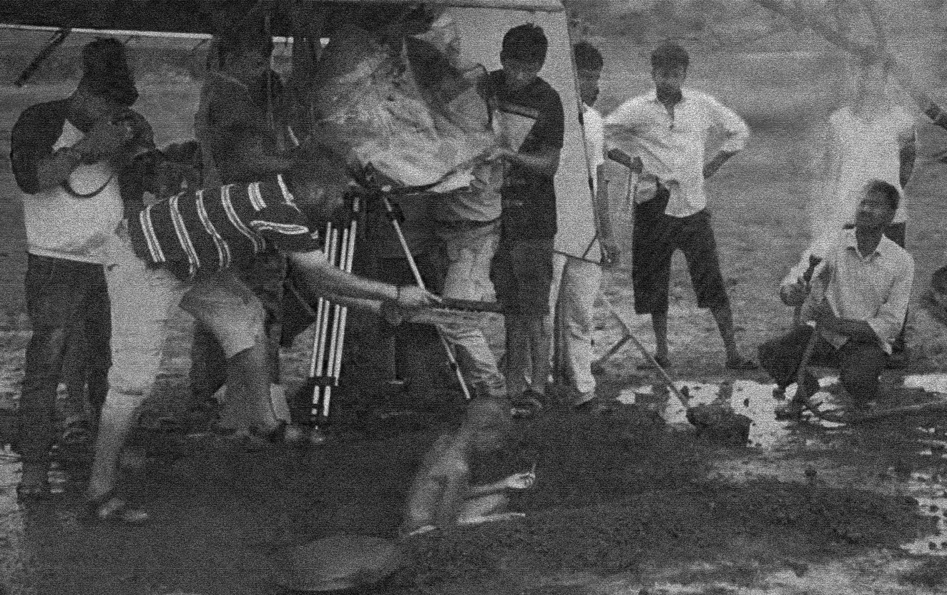 Black-and-white behind-the-scenes photo: a crew films with a camera on a tripod, sheltered under plastic sheeting, in a muddy village field as villagers look on.